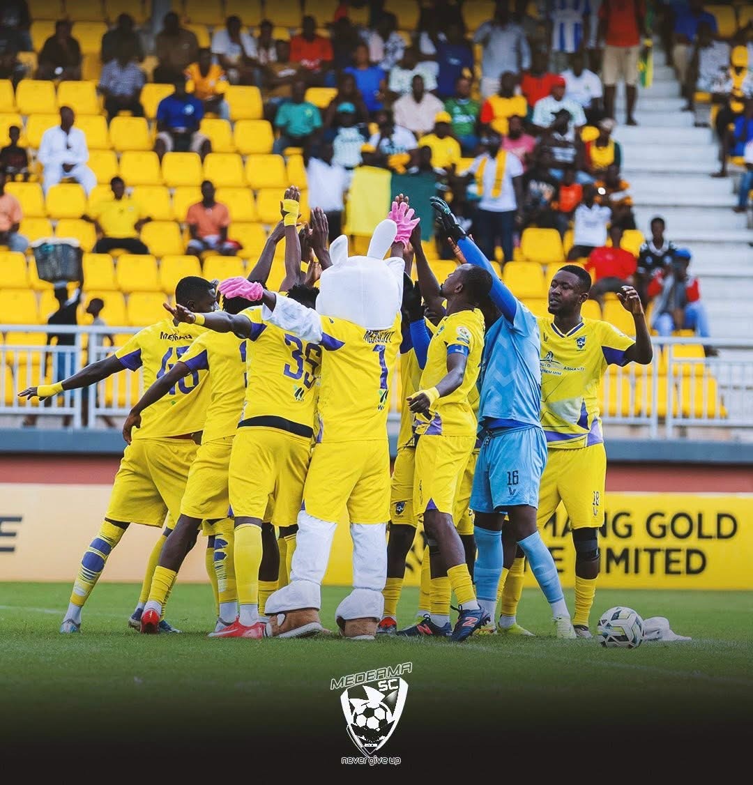 Soccer players in yellow uniforms celebrate a goal with a bunny mascot in the center, on a stadium field with yellow seats in the background.