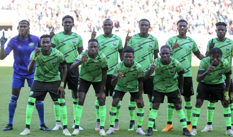 Soccer team posing for a group photo on the field, players in neon green kits with one finger raised, blue-jersey goalkeeper on the left.