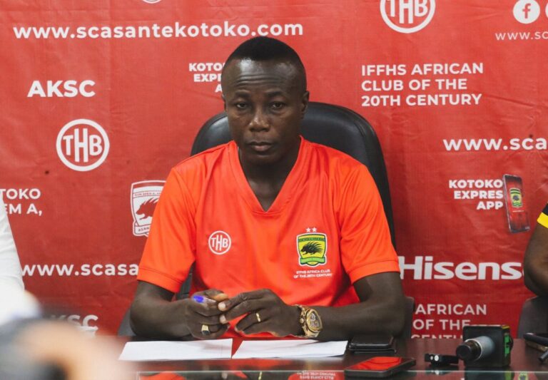 Man in an orange sports jersey at a press conference, seated with papers and a microphone in front, red sponsor backdrop behind him.