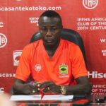 Man in an orange sports jersey sits at a table with papers, against a red banner backdrop full of logos and text like 'www.scasantekokoto.com'.