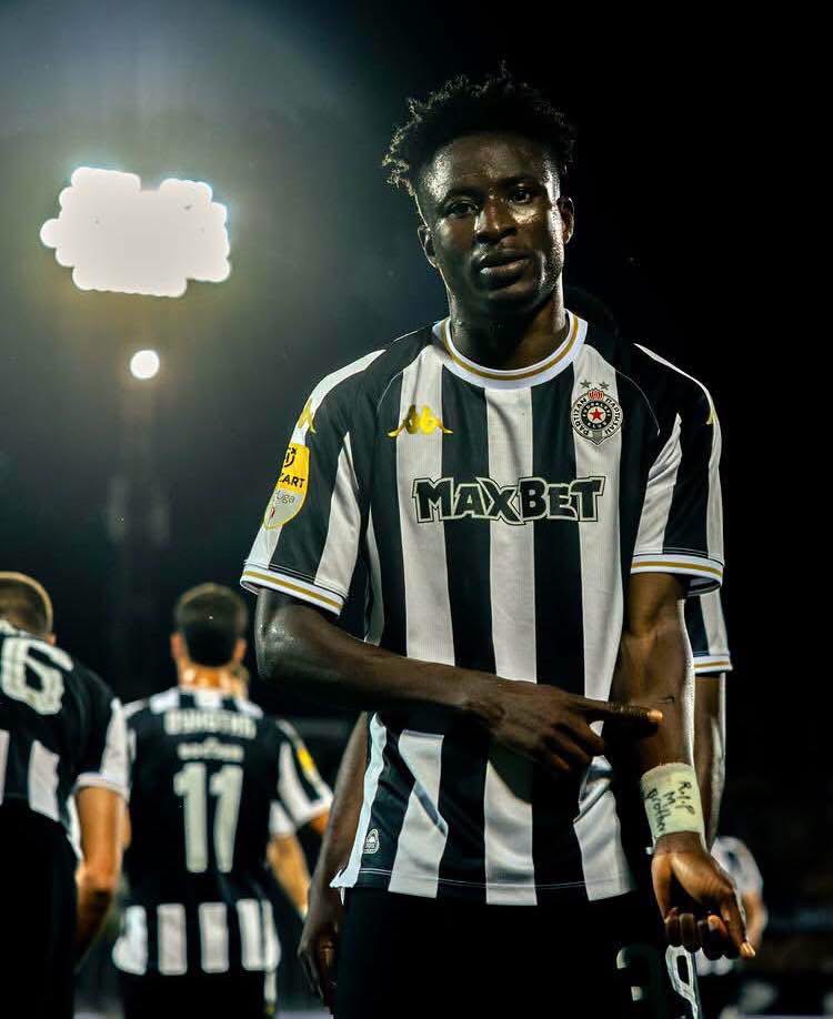 Soccer player in a black-and-white striped jersey with MAXBET sponsor, standing on a lit night field with teammates in the background.
