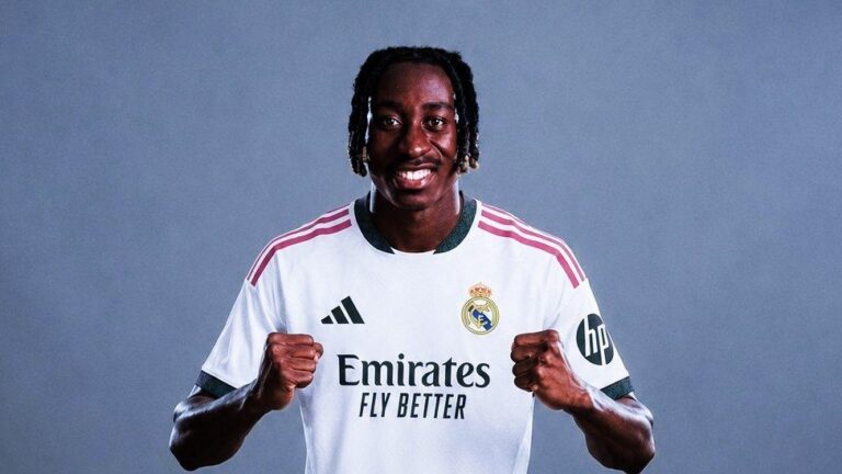 Soccer player in a Real Madrid jersey, smiling with fists raised in a celebratory pose against a plain blue-gray background.