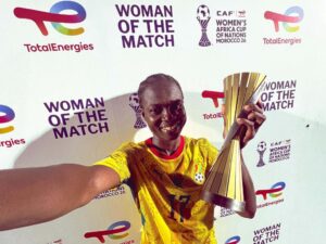 Female player in a yellow jersey holding a large trophy during a 'Woman of the Match' event, posing for a selfie against a backdrop with logos.