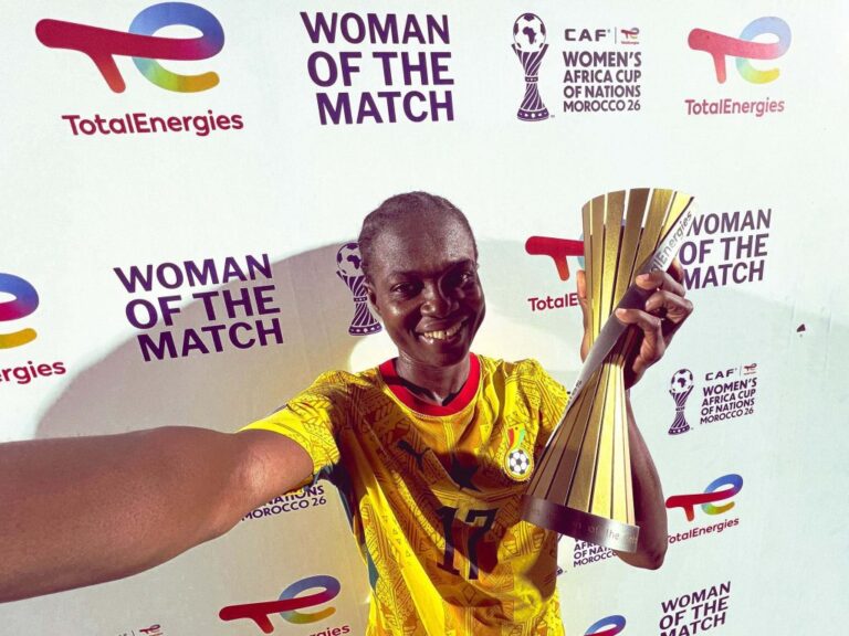 Female player in a yellow jersey holding a large trophy during a 'Woman of the Match' event, posing for a selfie against a backdrop with logos.