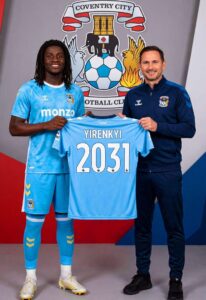 Two men stand side by side holding a light blue football jersey with the name 'YIRENKIYI' and number '2031' in front of a Coventry City crest backdrop, smiling.
