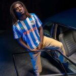 Young man with dreadlocks in a blue-and-white striped jersey stands beside an open car door in a dim setting, looking at the camera.