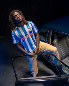 Young man with dreadlocks in a blue-and-white striped jersey stands beside an open car door in a dim setting, looking at the camera.