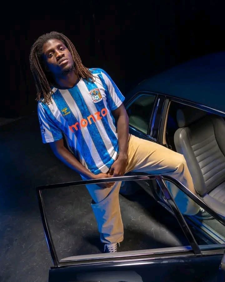 Young man with dreadlocks in a blue-and-white striped jersey stands beside an open car door in a dim setting, looking at the camera.