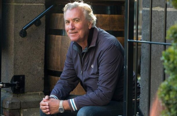 Older man with silver hair sits on stone steps outside a doorway, wearing a navy jacket and smiling at the camera.