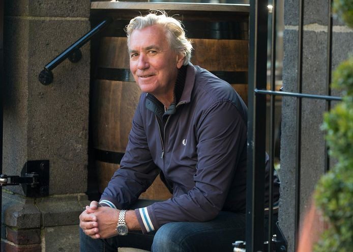 Older man with silver hair sits on stone steps outside a doorway, wearing a navy jacket and smiling at the camera.