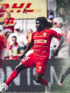 Soccer player in a red kit with number 16 kicking a ball during a match, with blurred spectators in the background.