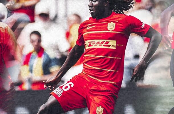 Soccer player in a red kit with number 16 kicking a ball during a match, with blurred spectators in the background.