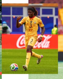 Soccer player in a yellow Ghana kit with number 3 dribbling the ball on a green field during a match, Coca‑Cola ad in the background