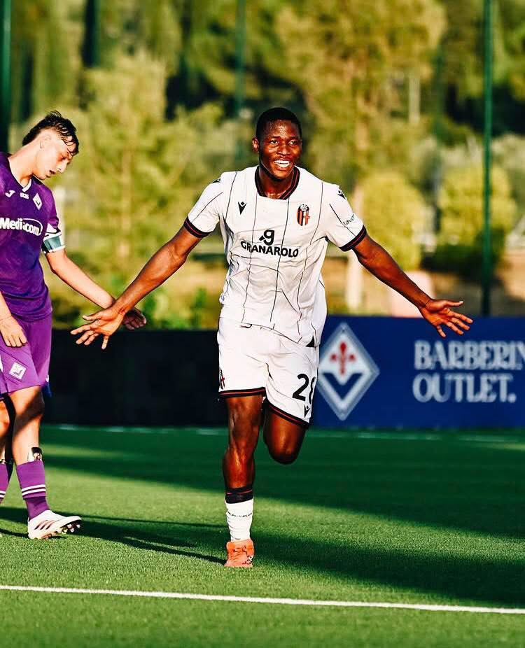 Soccer player in a white jersey with number 20 celebrates by running with arms outstretched on a sunny green field, a teammate in purple stands nearby.