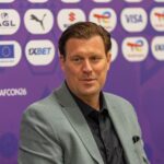 Man in a light gray suit and black shirt speaks at a press conference, microphone in front, purple sponsor backdrop behind him.