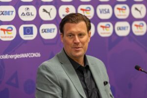 Man in a light gray suit and black shirt sits at a microphone during a press conference, with a purple sponsor backdrop behind him.