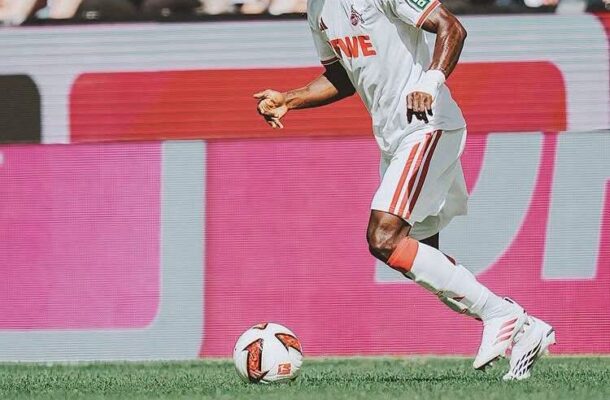 Soccer player in a white and red kit dribbling a ball on a green field with spectators in the background.