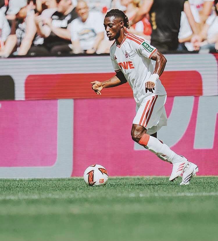 Soccer player in a white and red kit dribbling a ball on a green field with spectators in the background.