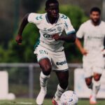 Soccer player in white kit dribbling a ball on the field, focused on the move with another player in the background
