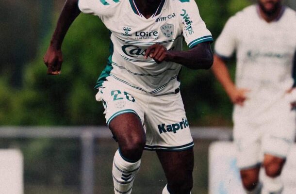 Soccer player in white kit dribbling a ball on the field, focused on the move with another player in the background