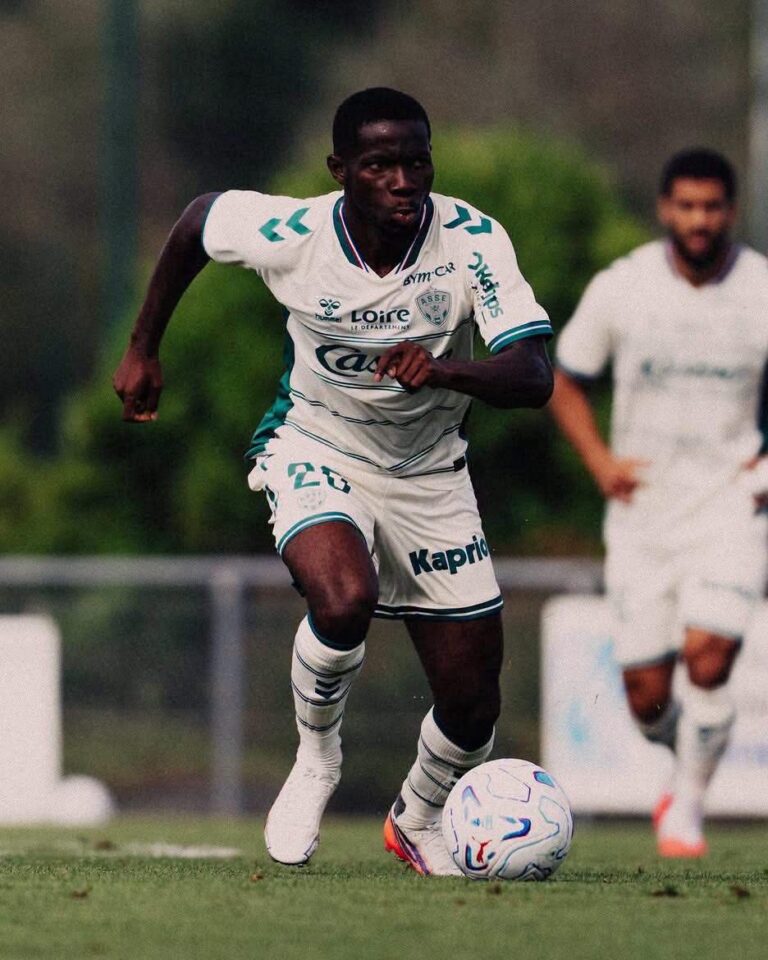 Soccer player in white kit dribbling a ball on the field, focused on the move with another player in the background