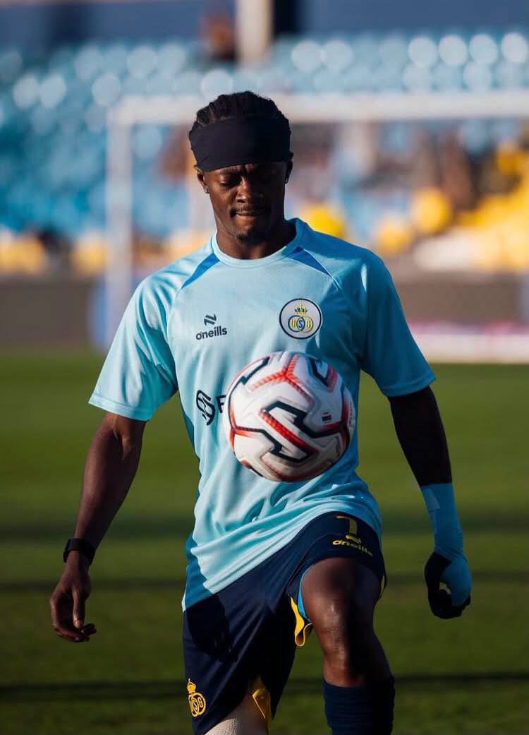 Soccer player in a light blue jersey traps a white and red football on a sunny field, stadium seats in the background.