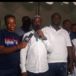 Man in white shirt speaks into a handheld microphone at a group event, flanked by people including a reporter with a TV mic and others in white attire.
