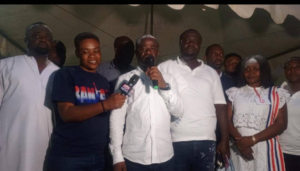 Man in white shirt speaks into a handheld microphone at a group event, flanked by people including a reporter with a TV mic and others in white attire.