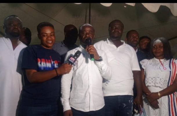 Man in white shirt speaks into a handheld microphone at a group event, flanked by people including a reporter with a TV mic and others in white attire.