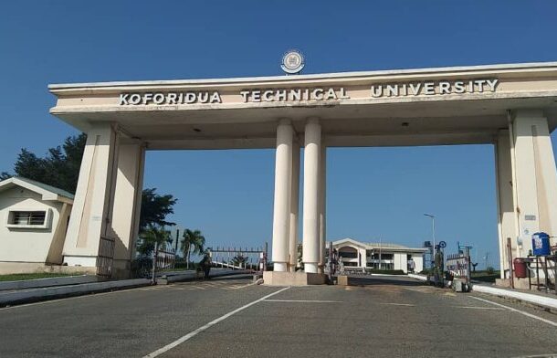 Entrance arch at Koforidua Technical University with tall white columns and a clear blue sky behind it, gate open to campus.