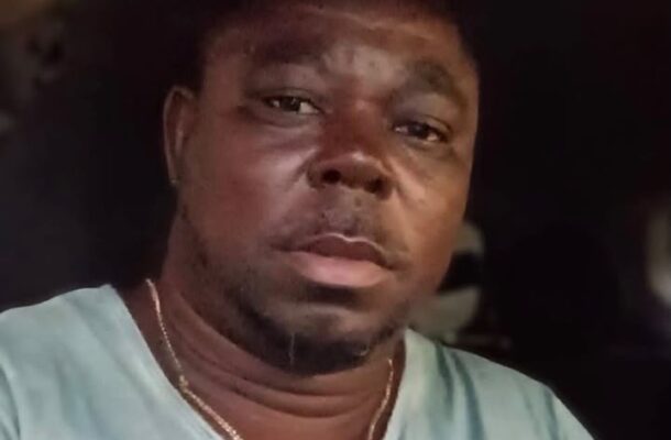 Close-up selfie of a man with short hair wearing a light blue shirt and a gold chain, inside a dimly lit vehicle, looking at the camera.