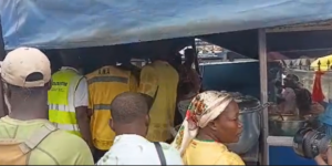 Crowd under a blue canopy at a market stall, with people wearing high‑visibility vests and casual clothing in line.