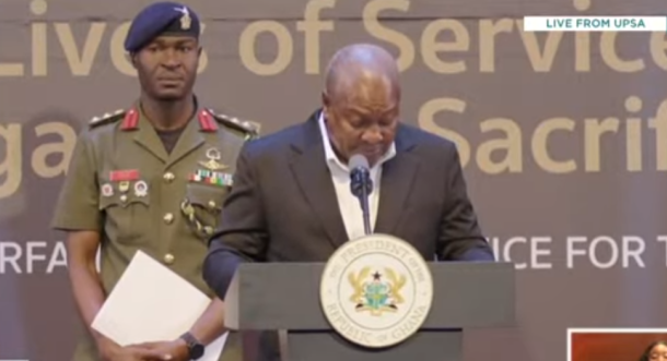 Man in a dark suit speaks at a podium with a presidential seal; a military officer in uniform stands behind holding papers, with a backdrop of large text and a 'Live from UPSA' banner.
