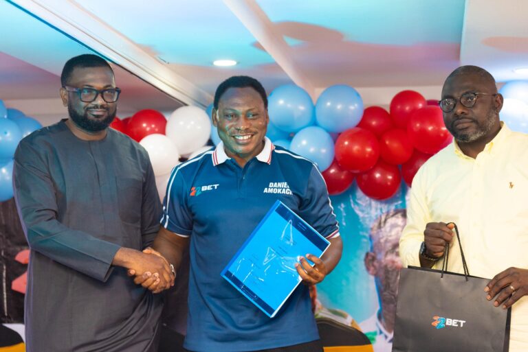 Three men in a celebration setting shake hands; one holds a blue certificate while balloons fill the background.