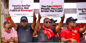 Protesters holding signs that read 'Courts Are for Justice, Not Political Revenge!' and 'You Can ARREST People. You Can't Arrest The Truth!' with a man wearing a red scarf in the center