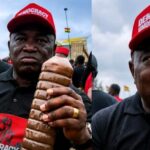 Two men at a protest wearing red caps that read 'DEMOCRACY UNDER ATTACK'; one is holding a large twisted brown beverage bottle with the crowd in the background.