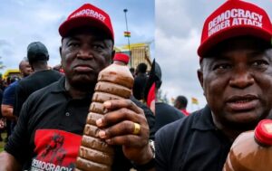 Two men at a protest wearing red caps that read 'DEMOCRACY UNDER ATTACK'; one is holding a large twisted brown beverage bottle with the crowd in the background.