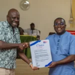 Two men shake hands while one presents a certificate at a formal event, both smiling.