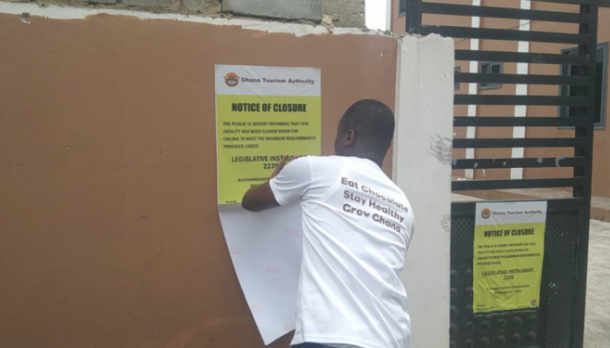 Man in a white T‑shirt posts a yellow 'Notice of Closure' sign on a brown wall beside a gated area.