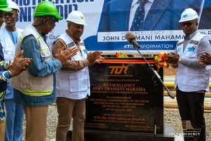 Construction workers and officials unveil a commemorative plaque at a ceremony, with a banner featuring John Dramani Mahama in the background.