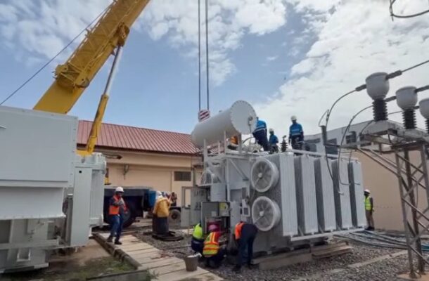 Large electrical transformer being moved into position by a yellow crane, workers in safety vests and helmets nearby at a substation site.