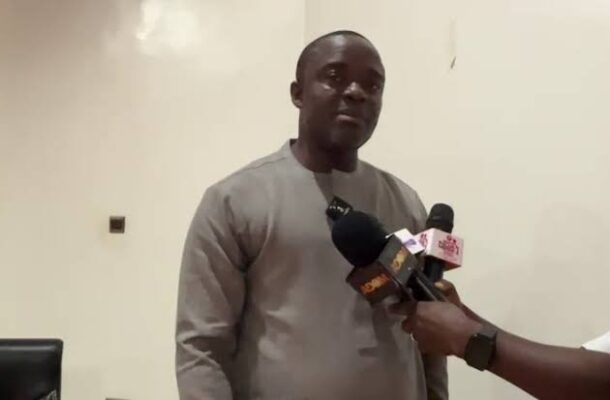 images | Ghanaguardian.com Man in a gray traditional outfit being interviewed by reporters, several microphones held toward him in a room.