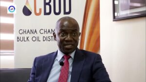 Man in a navy suit and pink tie speaks in an interview, microphone clipped to his lapel, with a banner reading 'GHANA CHAMBER BULK OIL DIST' behind him
