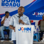 Male speaker at a campaign event delivering a speech from a podium labeled 'JOHN BOADU' on a blue stage with supporters seated behind him.