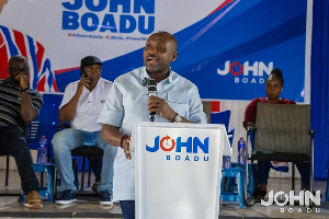 Male speaker at a campaign event delivering a speech from a podium labeled 'JOHN BOADU' on a blue stage with supporters seated behind him.