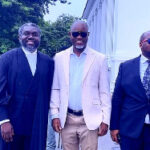Three men in suits posing together at an outdoor formal event, with trees and a white building in the background.