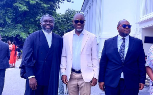 Three men in suits posing together at an outdoor formal event, with trees and a white building in the background.