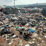 Open-air garbage dump with scattered trash across the ground and people in the background under umbrellas.