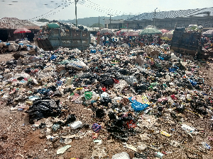 Open-air garbage dump with scattered trash across the ground and people in the background under umbrellas.