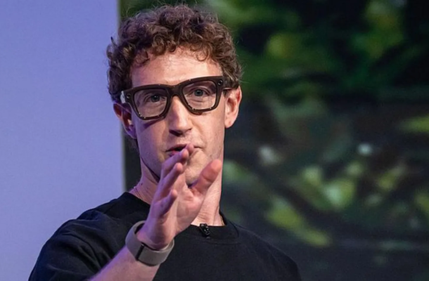 Speaker with curly hair and dark glasses speaks on stage, gesturing with his right hand.